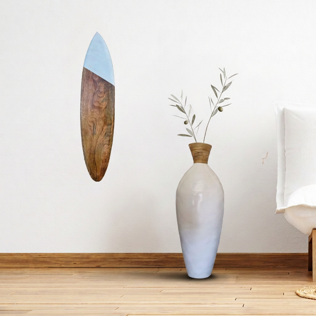 Wooden surfboard with a pale blue tip and visible natural grain hanging beside a tall white floor vase with sparse olive branches.