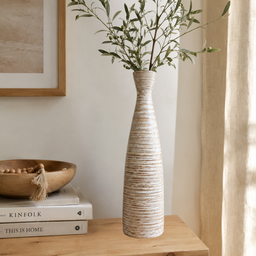 A tall, narrow ceramic vase with a textured, horizontally ribbed beige and white finish, holding green branches, styled on a wooden table next to a bowl and books in a warm, neutral-toned room.