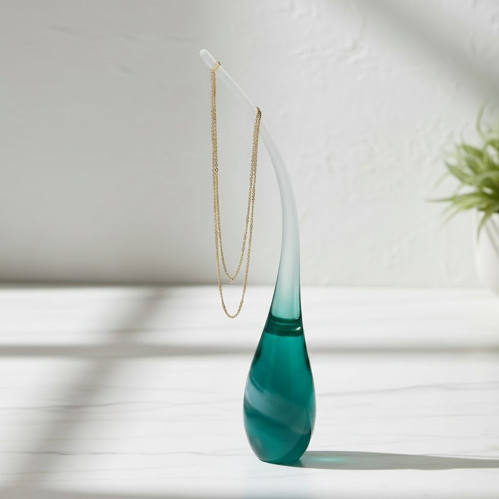 Teal glass sculpture with frosted curved stem, gold chain necklace draped over the stem and gold bangles resting at the base, soft shadows on a white surface with a green plant in the background

