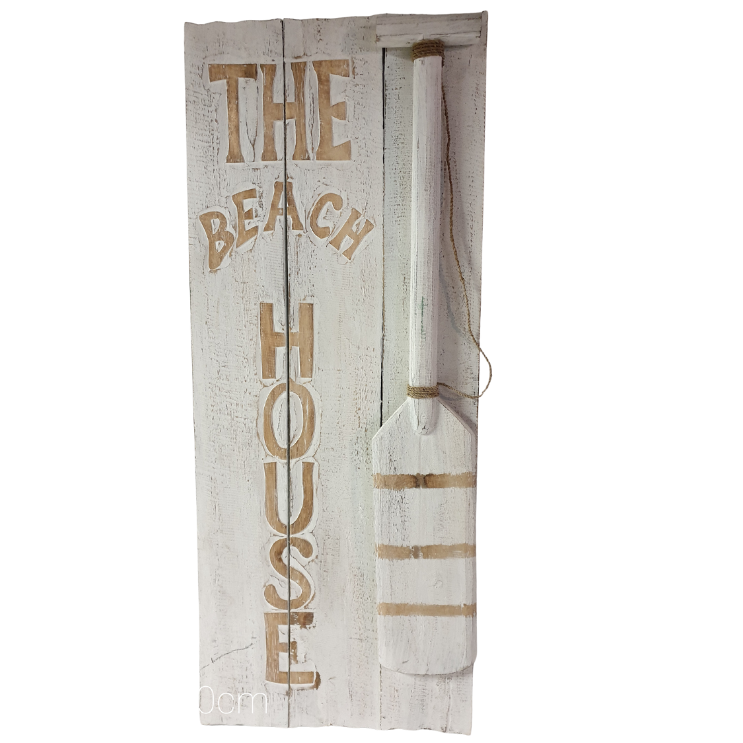 A rustic vertical wooden wall sign featuring distressed white wood planks with the words 'THE BEACH HOUSE' in a tan stencil font and a matching white paddle illustration on the right side.