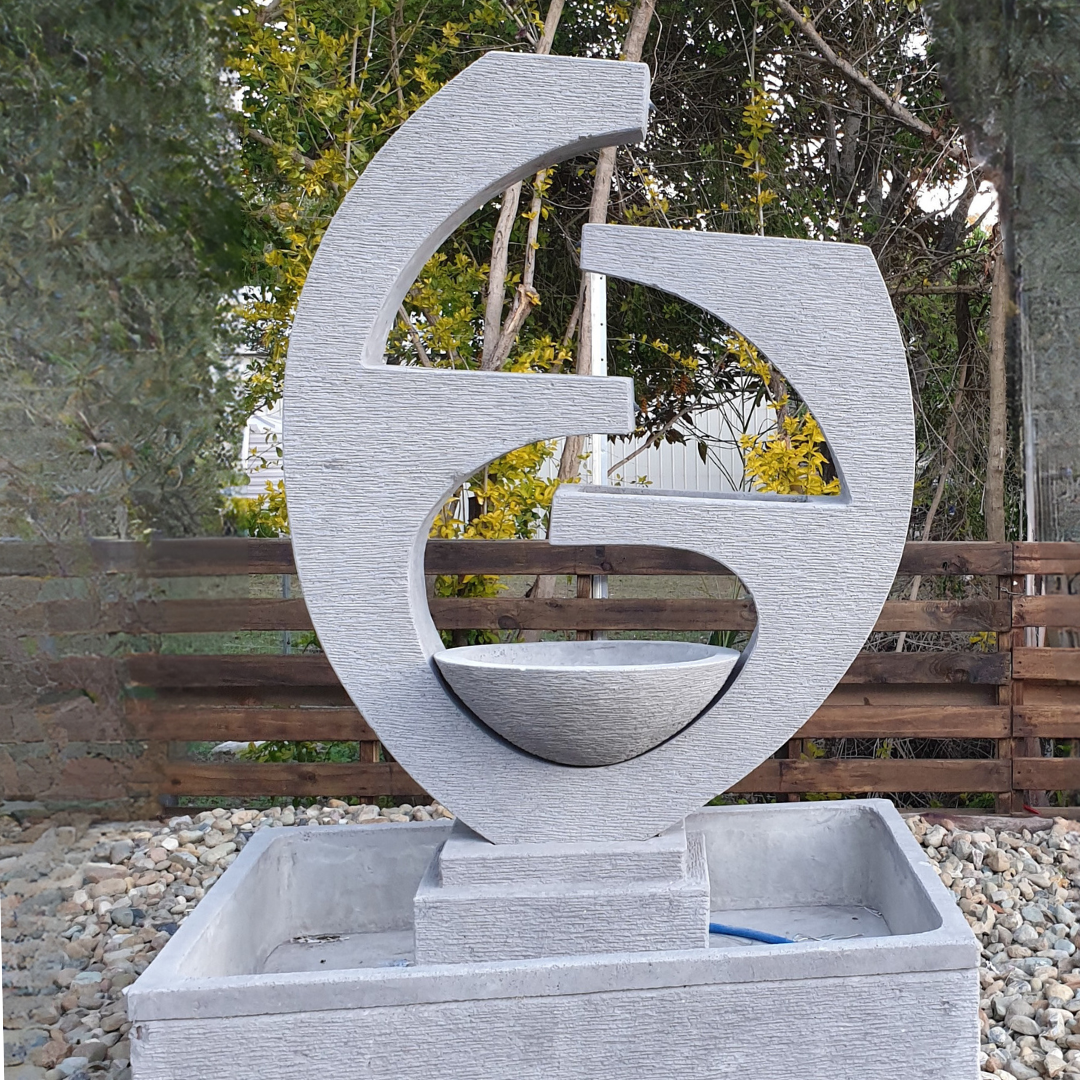Sculptural concrete garden fountain as a focal point in a landscaped outdoor setting with timber bench and garden chairs on stone paving