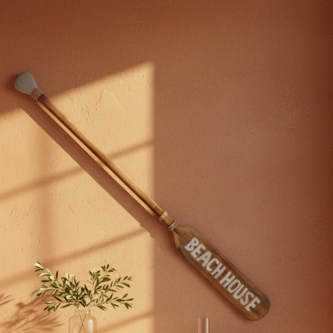 A wooden paddle-style wall decor piece with the words 'BEACH HOUSE' painted on the blade, mounted on a warm terracotta wall with sunlight casting shadows and a small plant in the lower left.
