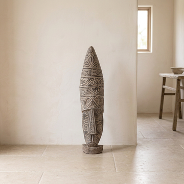 Whitewashed wooden tribal mask with carved spiral and geometric patterns on a round base, displayed on a pale stone floor in a neutral interior.