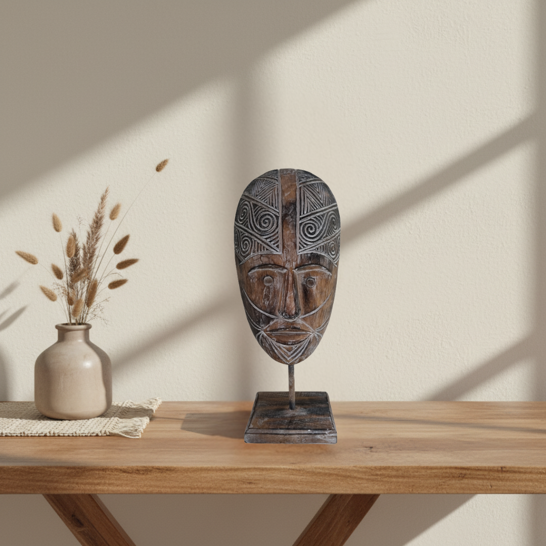 A carved wooden African-style mask sculpture on a metal stand, displayed on a wooden table next to a small beige vase with dried grass against a warm beige wall with soft window shadows.