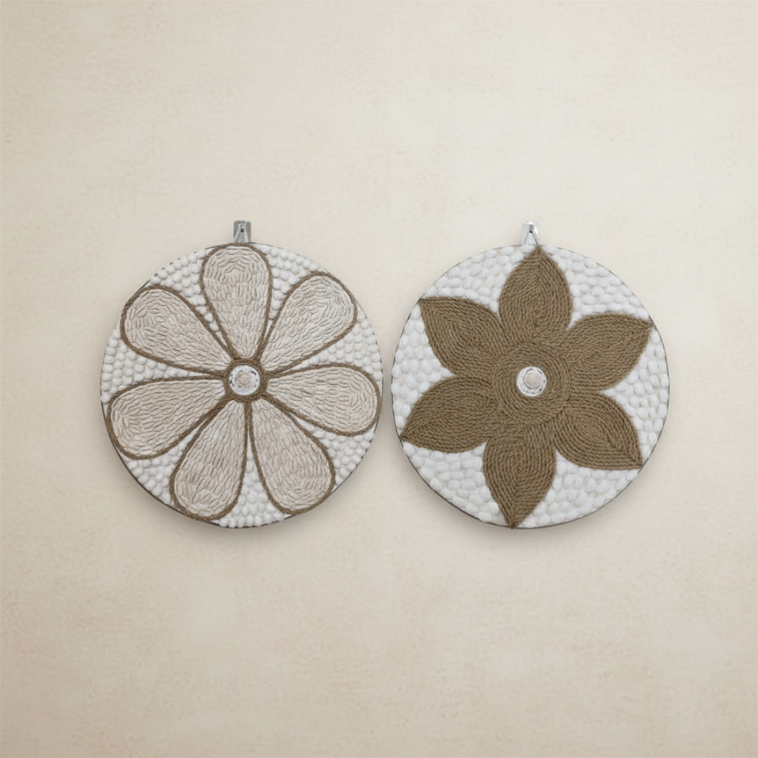 Two round handcrafted wall art pieces laid flat on a warm beige surface — the left with a soft cream jute daisy petal pattern and the right with a bold tan jute star flower, each with a central shell accent.