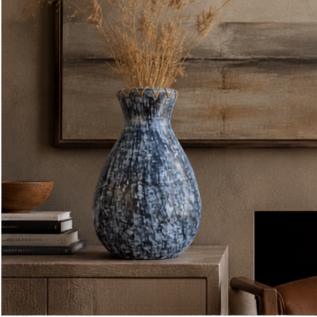 Teardrop-shaped pot with dark blue-grey distressed finish and raised oval dots, woven jute collar at the opening, filled with dried wheat grass, styled on a timber sideboard with stacked books and a wooden bowl, large framed artwork on the wall behind