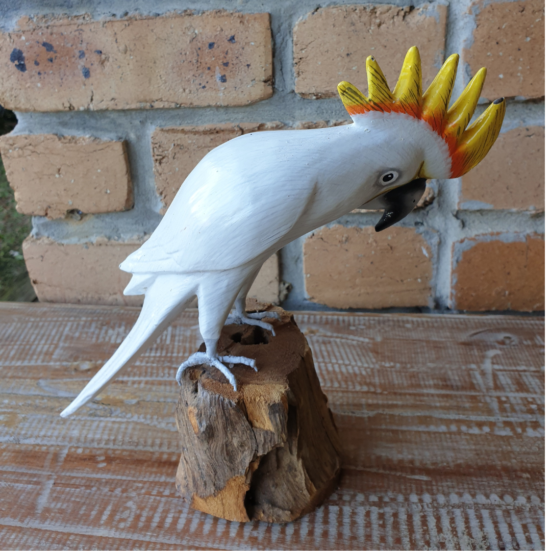Model of a white cockatoo with a yellow crest on a wooden perch against a brick wall.