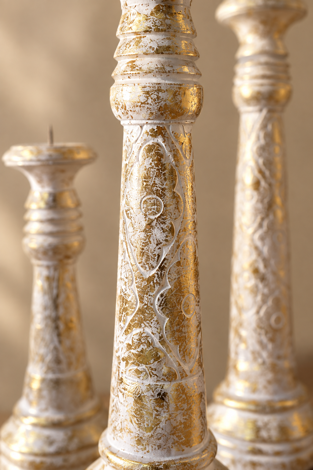 Close-up of ornate candlesticks with a distressed gold and white finish, showing carved decorative details on the central candlestick against a softly blurred beige background.
