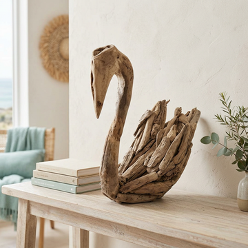 Natural driftwood bird with a curved neck and layered wooden wings on a pale timber console beside stacked books and a vase of greenery.