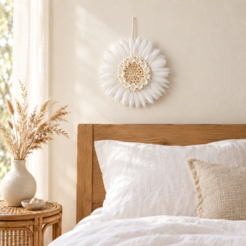 White feather and shell wall hanging above a timber bedhead with white linen bedding and a rattan side table.
