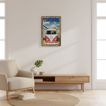 A framed vintage-style travel poster reading 'Gone Surfing' hangs on a white wall above a light wood console table in a bright, modern living room with a beige armchair and woven rug.