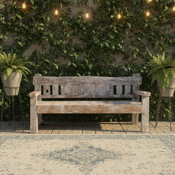 Weathered white-painted bench seat with slatted backrest and chunky armrests, centred on a patterned outdoor rug beneath warm string lights against an ivy-covered wall with potted ferns