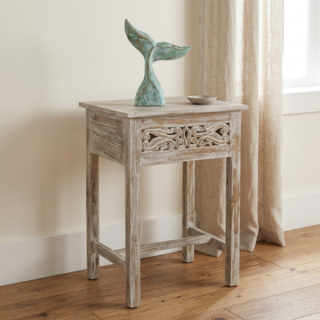 Distressed turquoise ornament with curved flukes displayed on a whitewashed carved side table beside a sunlit window