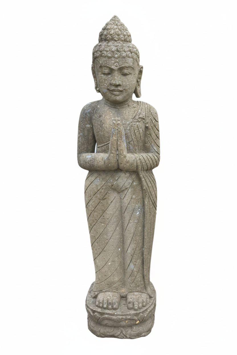 Standing hand-carved stone Buddha statue with hands pressed together in namaste prayer gesture, dark olive-grey weathered stone with rough texture, on a lotus base, white background