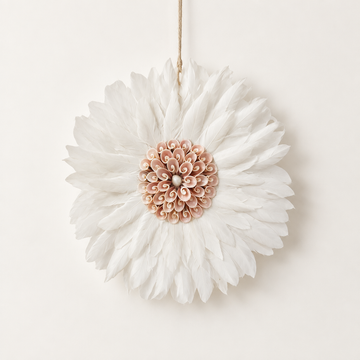 White JUJU hat wall art with layered white feathers radiating around a blush pink spiral shell rosette centre, hanging from a natural cord.