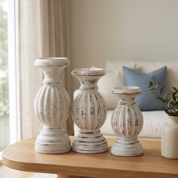 Three distressed white carved stands on a rounded timber console, with a cream vase and blue sofa cushion behind.